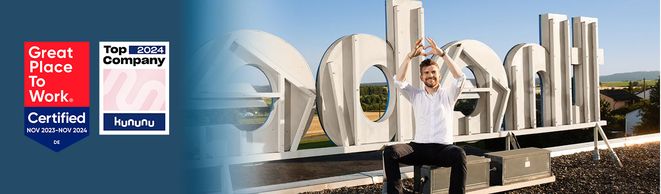 Theben-Geschäftsführer Paul Sebastian Schwenk zeigt mit Händen ein Herz vor dem Theben-Firmenlogo auf dem Büro-Dach, daneben Siegel „Great Place to Work- Zertifiziert“ und „Kununu Top Company 2024"