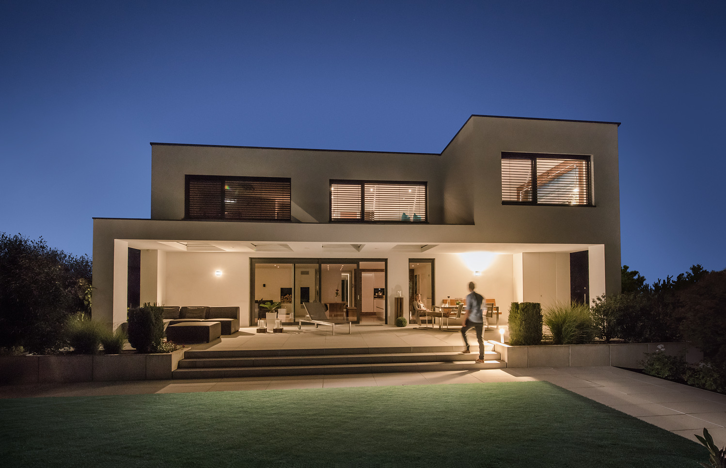 Moderne Hausfassade bei Nacht mit beleuchteter Terrasse, Außenbeleuchtung und Person in Bewegung