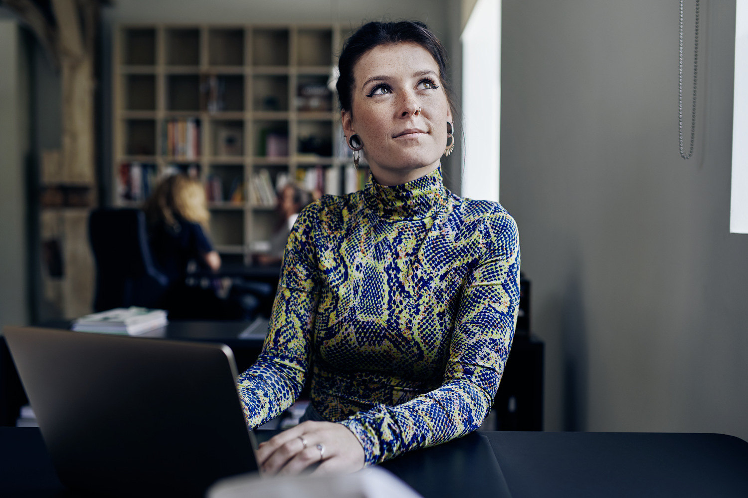 Nachdenkliche Frau an einem Laptop in modernem Arbeitsumfeld mit Bücherregal im Hintergrund